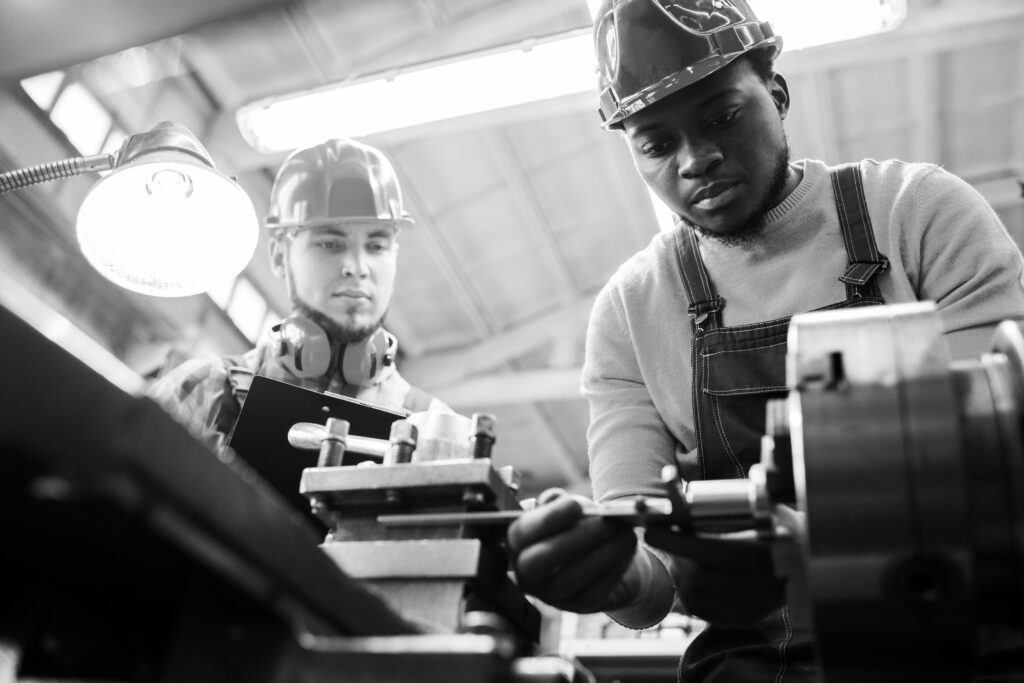 Serious concentrated African-American factory engineer in hardhat adjusting milling machine and joining detail while skilled inspector examining his work and controlling production process