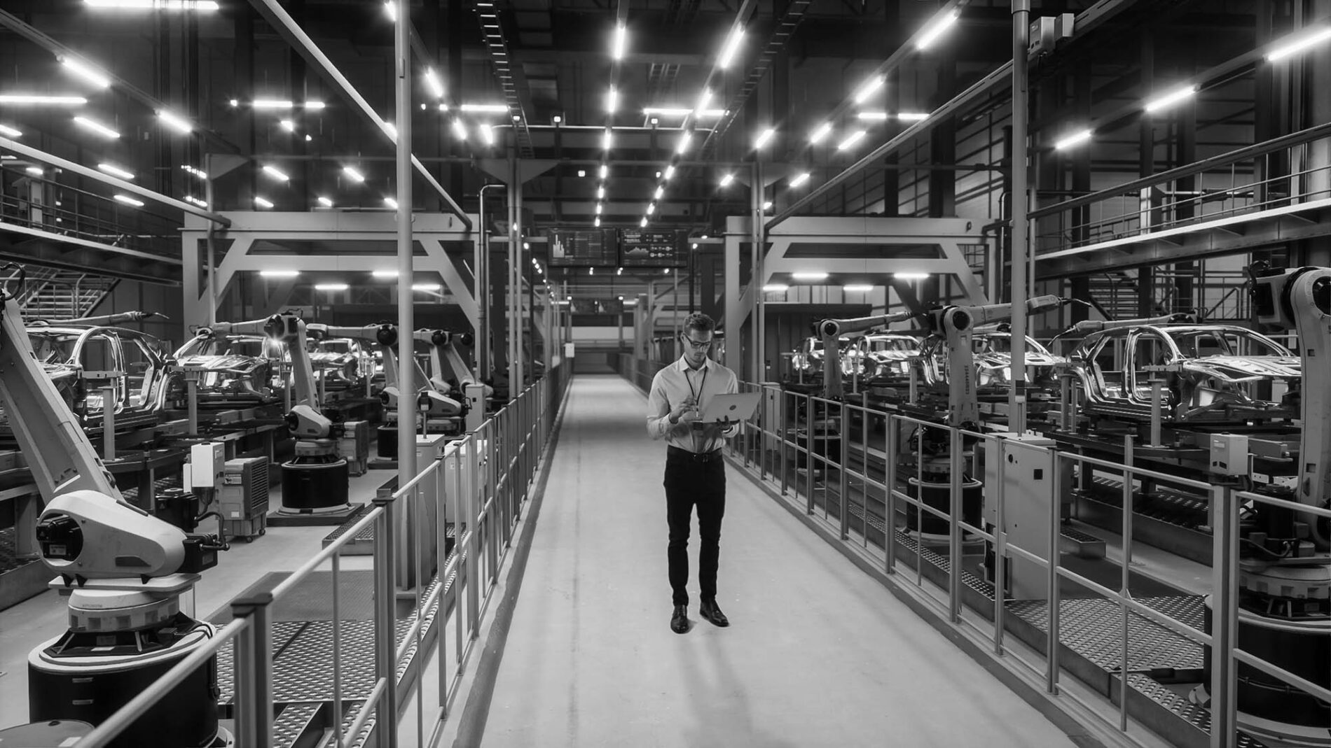 Engineer Holds Laptop Walks through Autonomous Automotive Assembly Line. Robotic Arms and Car Frames. Industrial Manufacturing Facility Working on EV Vehicle Production. Automated Assembly Plant.